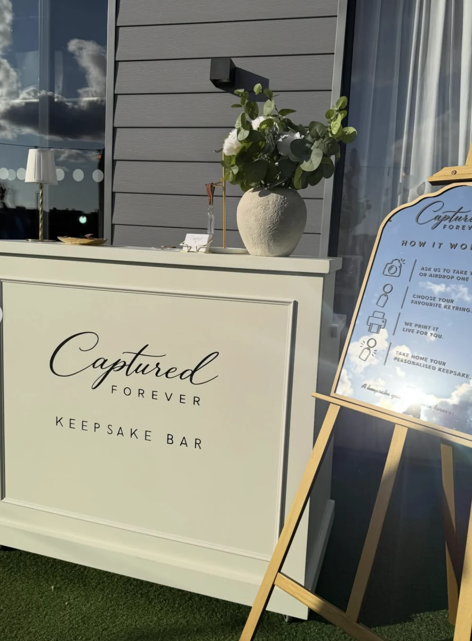 The Captured Forever Keepsake Bar and mirrored How It Works easel set up on a lawn at an evening event