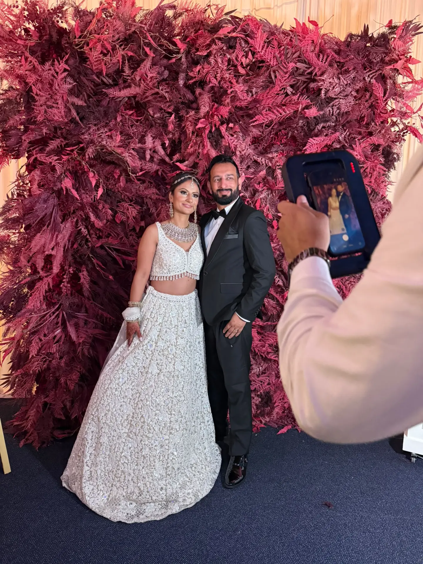 A couple being photographed in front of a deep red foliage wall at their wedding reception