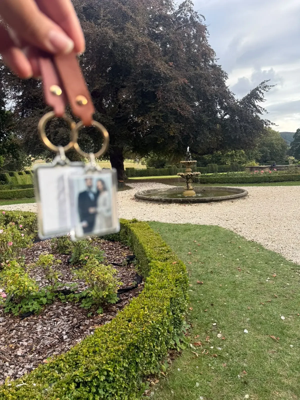A pair of finished personalised photo keyrings held in front of a formal garden fountain
