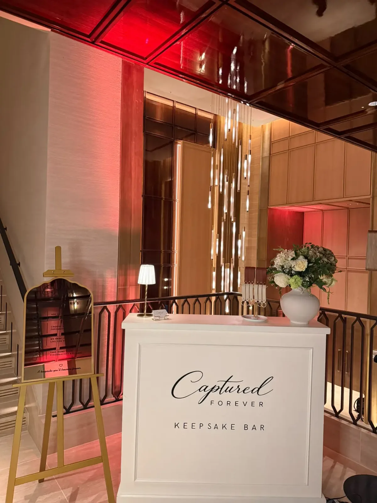 The Captured Forever Keepsake Bar and mirrored How It Works easel styled with white roses in a warmly lit hotel interior